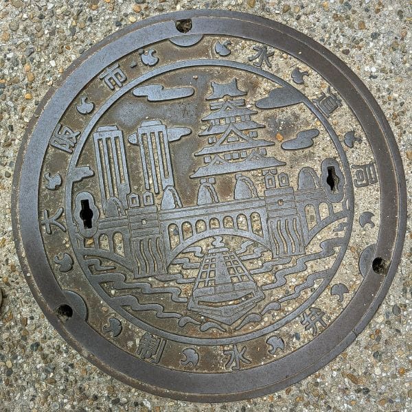 Decorative manhole cover in Osaka featuring an engraved design of Osaka Castle, a bridge, and city landmarks.