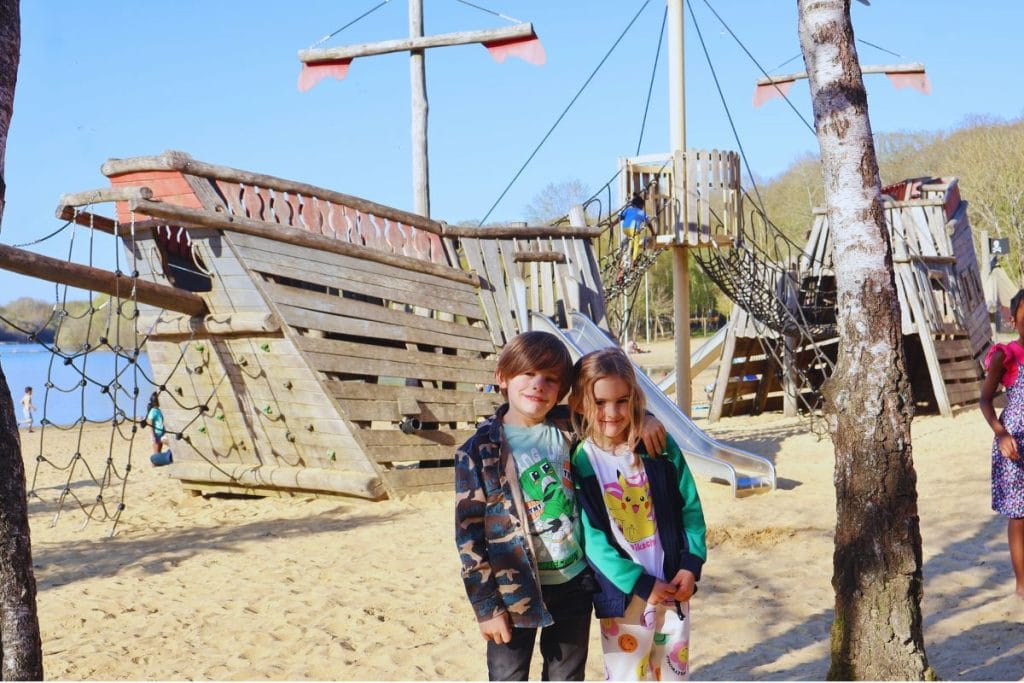 “Children standing in front of the wooden pirate ship playground at Ruislip Lido, set in sand with climbing nets and slides, on a sunny day.”