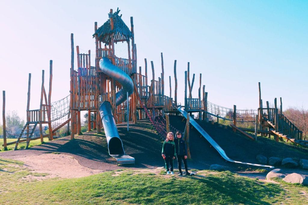 Two children standing in front of the large wooden adventure playground at Northala Fields in Northolt, featuring tall rope bridges and twisty tube slides under a clear blue sky.”