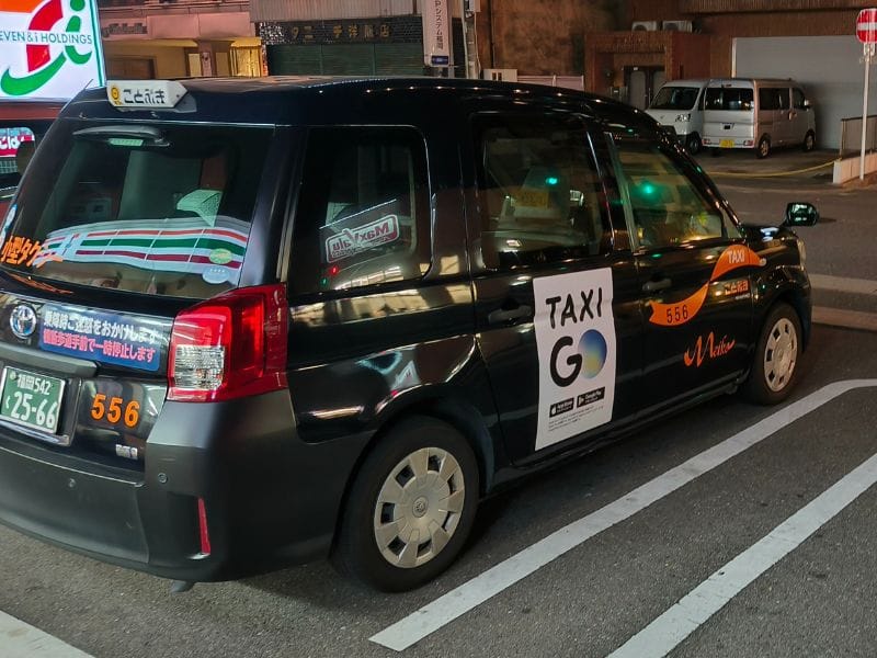 Photo of a Japanese taxi with the Go Taxi app logo and branding, parked near a 7-Eleven in Osaka at night