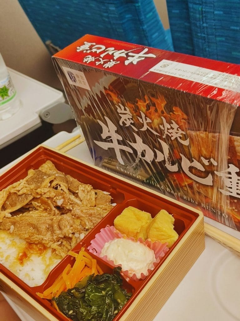Japanese bento box meal with grilled beef, rice, and side dishes, served on a tray table aboard the Shinkansen between Osaka and Fukuoka.