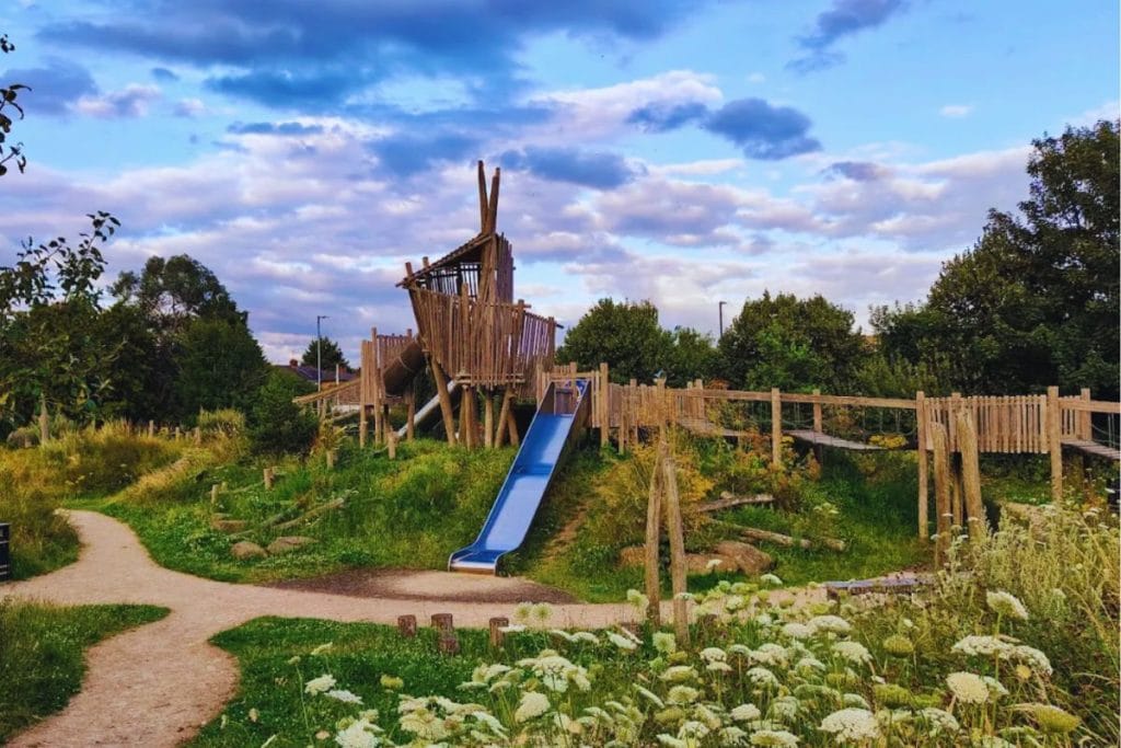 “Wooden adventure playground at Harwoods in Watford, featuring rope bridges, a tube slide, and a bright blue slide surrounded by wildflowers under a dramatic cloudy sky.”