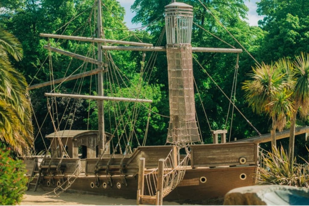 Large wooden pirate ship climbing structure at the Princess Diana Memorial Playground in Kensington Gardens, surrounded by palm trees and greenery under a bright blue sky.
