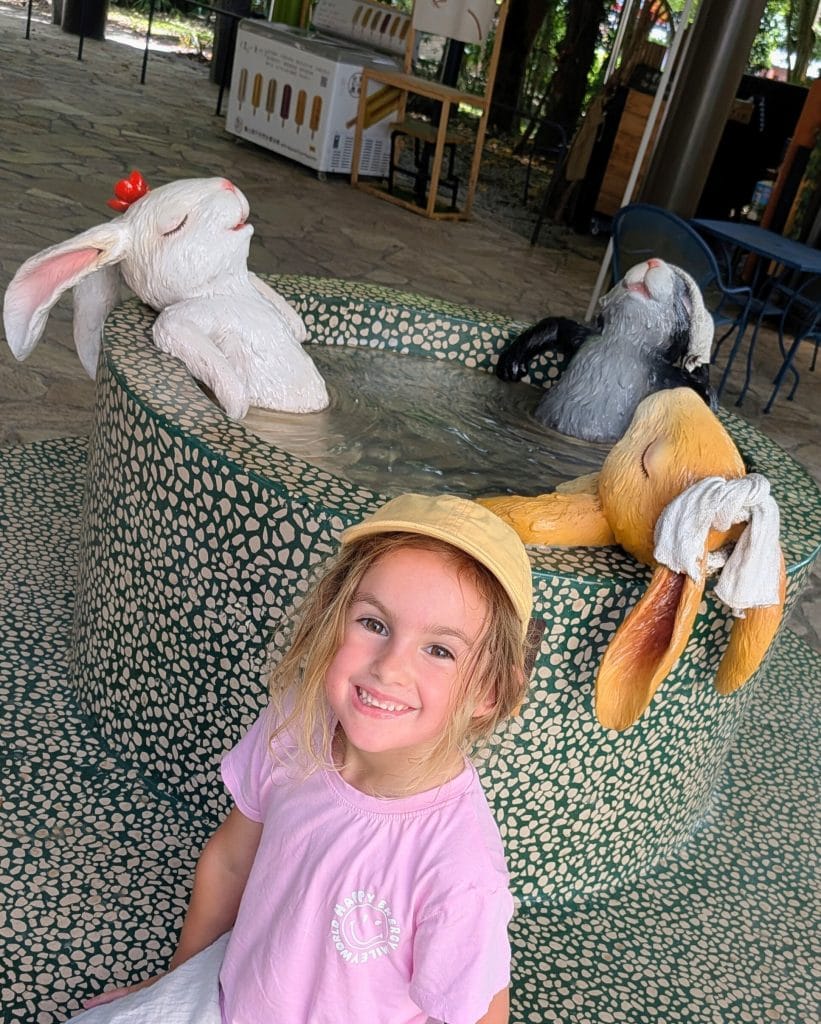 Smiling young girl at Forrest Bath Hot Springs in Jaoxski, posing in front of whimsical rabbit sculptures soaking in a tiled hot spring pool