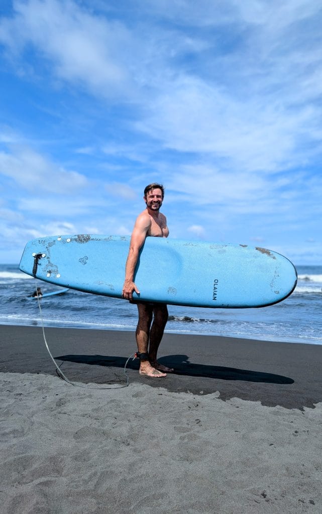 Man holding a blue surfboard on the black sand shores of Wai’ao Beach, Yilan, Taiwan—perfect spot for beginner-friendly surfing with scenic coastal views.