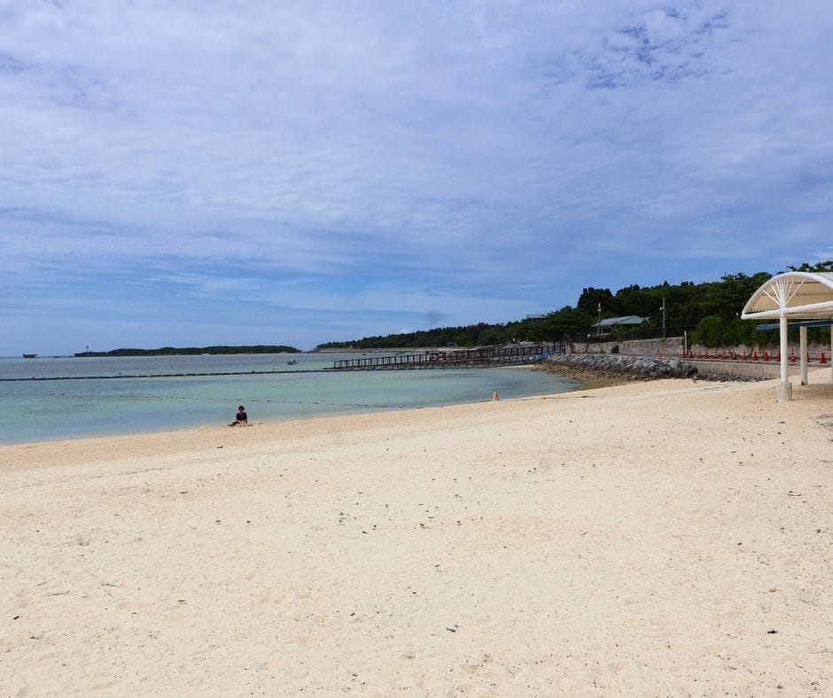 Child sitting on the sand at Emerald Beach in Okinawa with calm turquoise water and a long pier in the background.