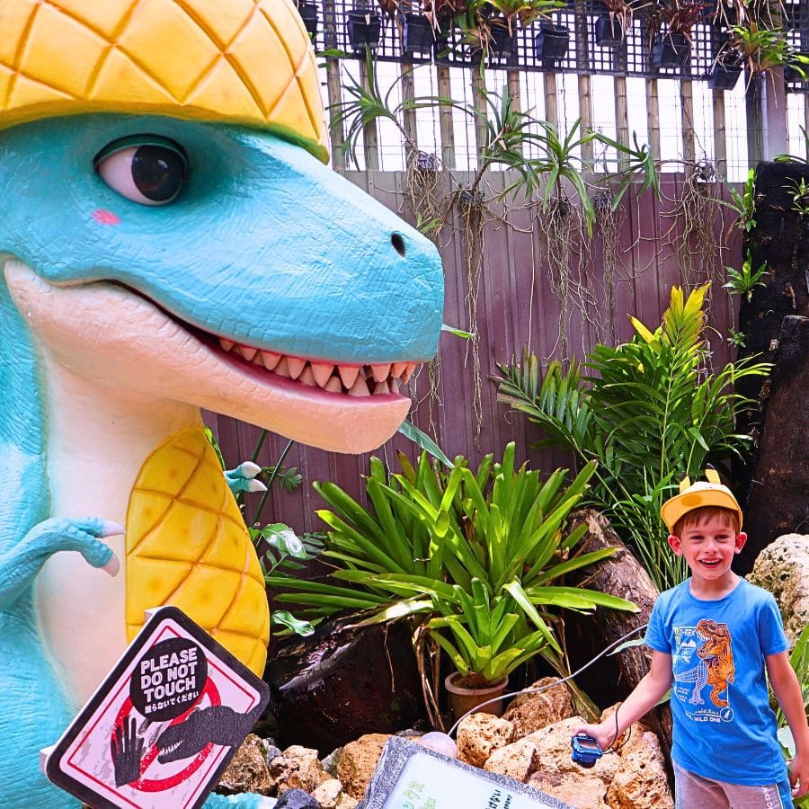 Child smiling next to a pineapple-themed dinosaur statue in the Dinosaur Zone at Nago Pineapple Park, Okinawa