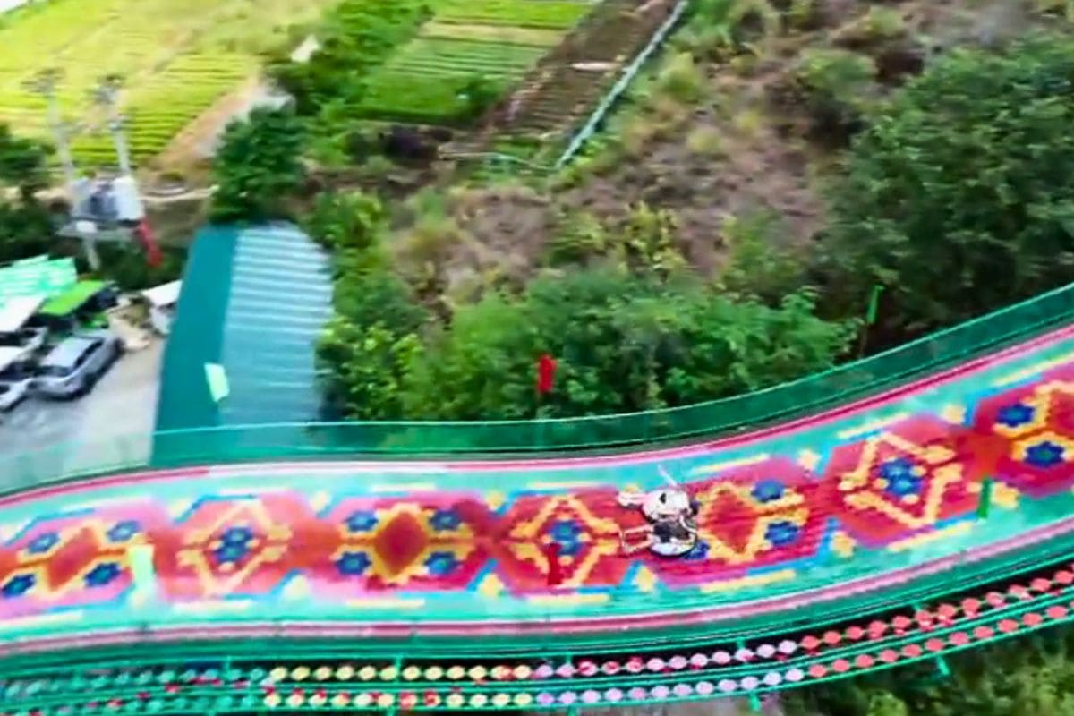 "Two people riding together on a sled down Sapa’s vibrant rainbow slide, decorated with bold geometric patterns, with green hills and farmland in the background."
