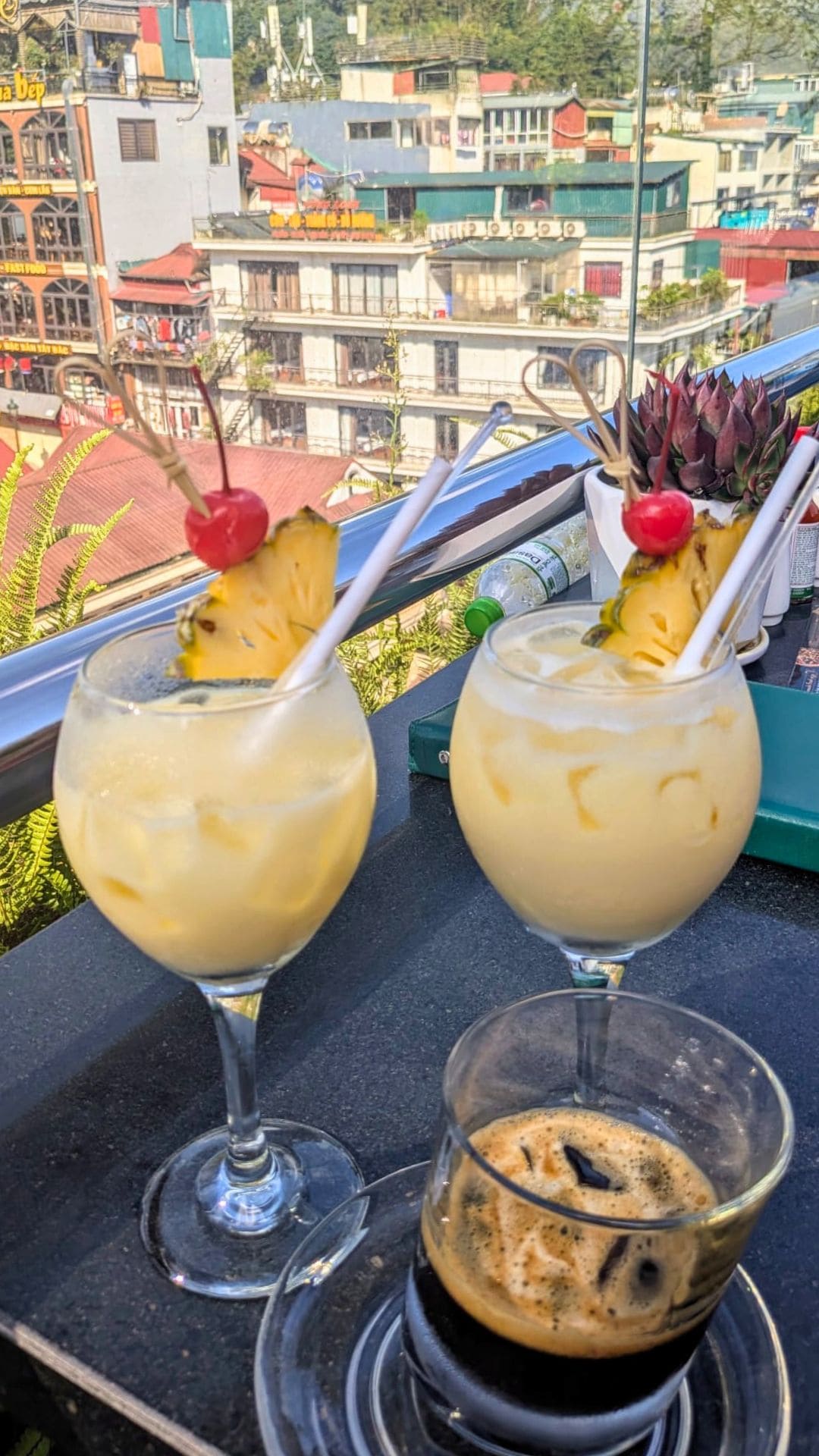 "Tropical cocktails with pineapple and cherry garnish alongside an espresso on a rooftop bar table, overlooking the colorful buildings of Sapa, Vietnam"