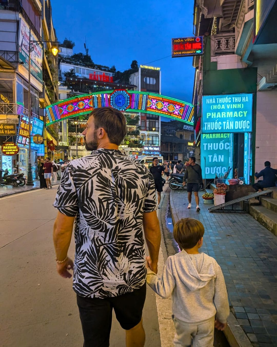 Father and son walking along Sapa’s main high street in Vietnam, surrounded by shops and mountain scenery — family travel
